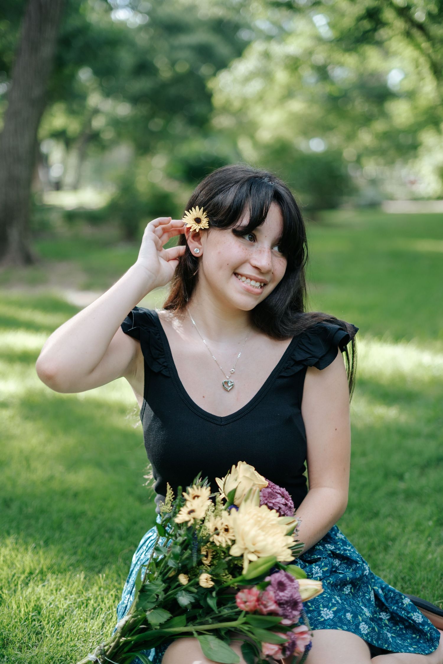 Smiling young woman holding colorful bouquet in park, tucking yellow flower behind ear, wearing black top and floral skirt.