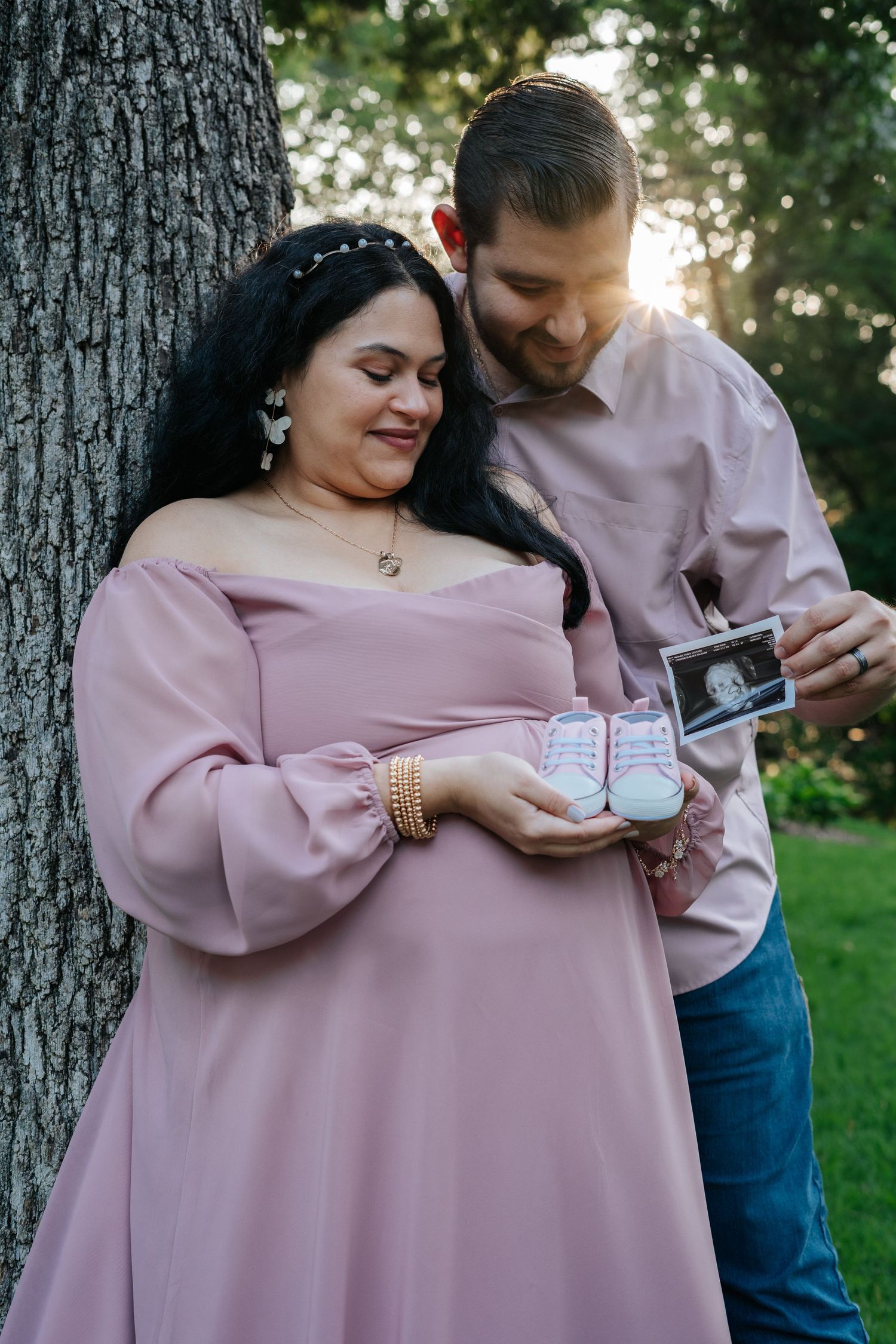 Pregnant couple holding baby shoes and ultrasound photo by a tree in golden hour light.