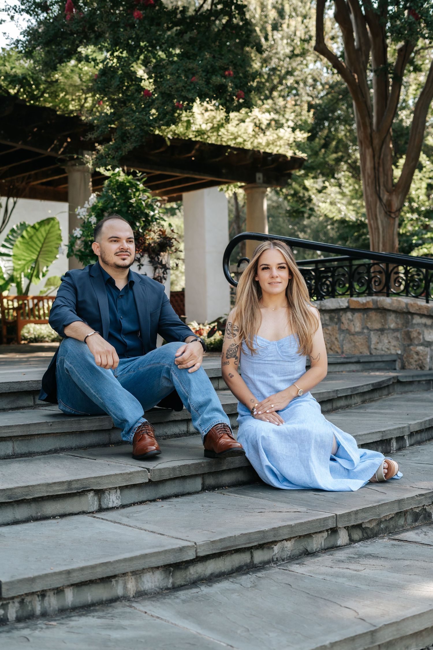 Couple sitting on stone steps in a garden, man in navy blazer and jeans, woman in light blue dress, lush greenery behind.