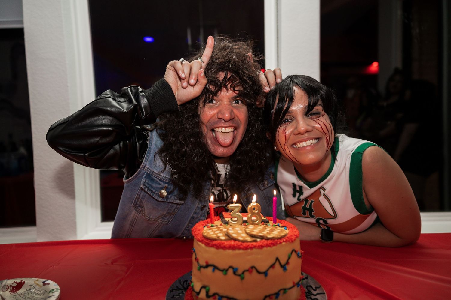 Two people in costumes celebrate with a decorated birthday cake with lit candles at a party.
