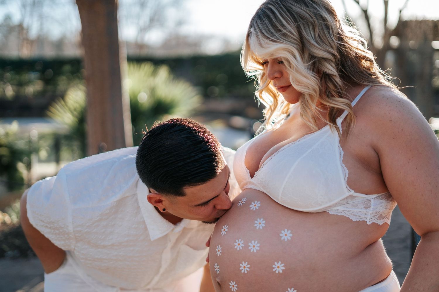 Couple sharing a tender maternity photo moment outdoors, man kissing pregnant woman's belly with floral designs.