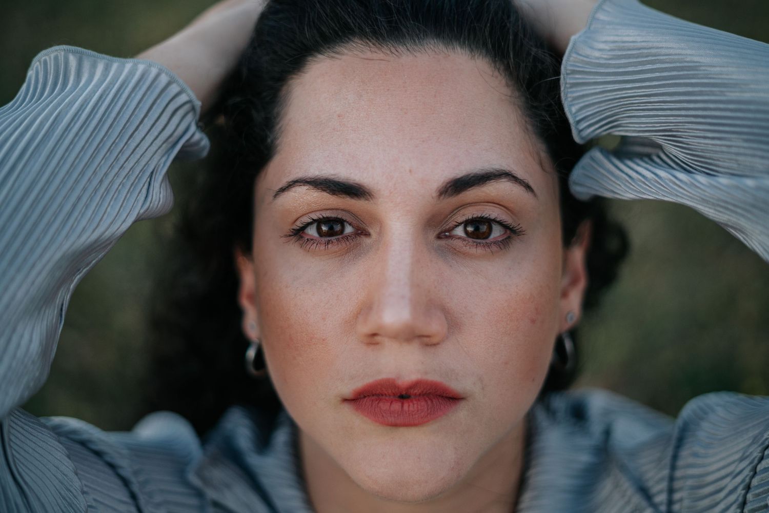 Close up portrait of a young woman with dark curly hair, freckles, and hoop earrings, hands raised behind her head.