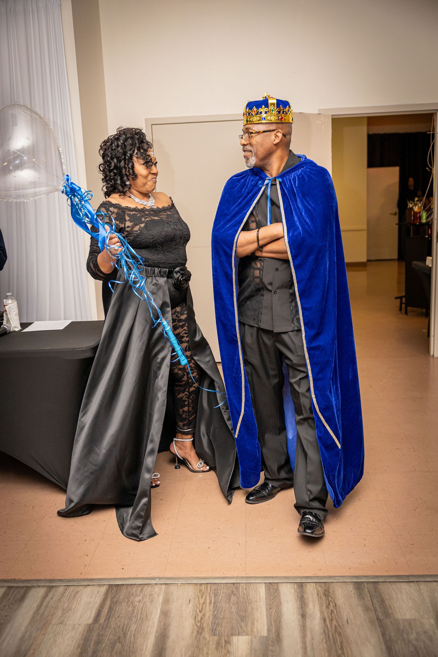 Two performers in blue velvet cape and black dress dance together in an elegant ballroom setting.