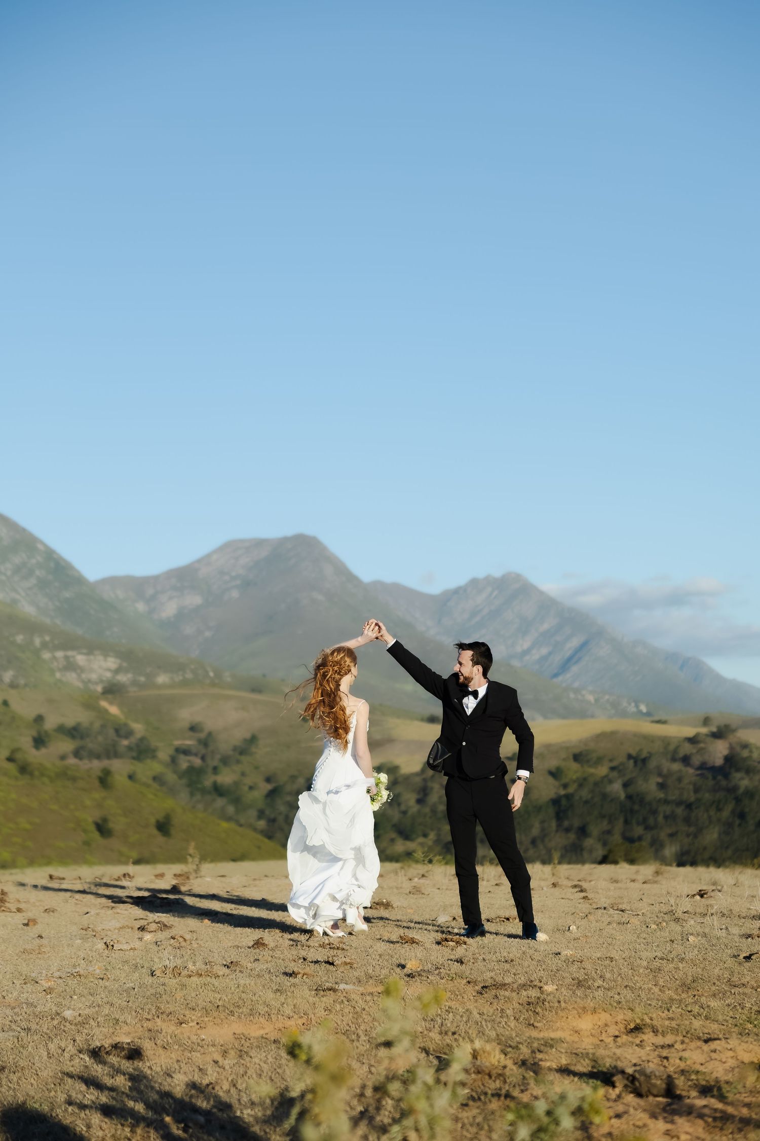 A couple in wedding attire runs playfully across a scenic mountain landscape under bright blue skies.