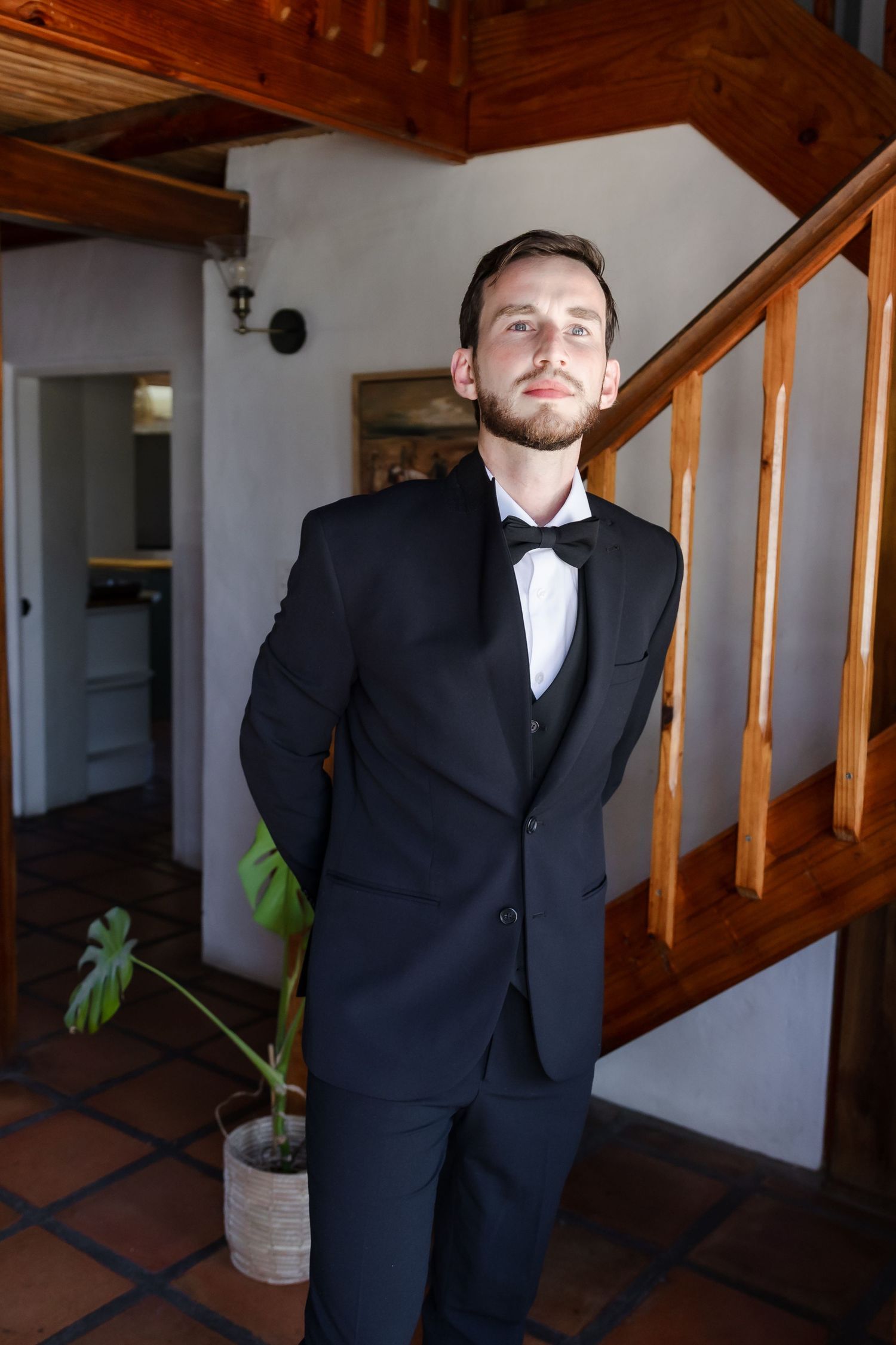 A formal portrait of someone in a classic black tuxedo and bowtie standing by a wooden staircase.