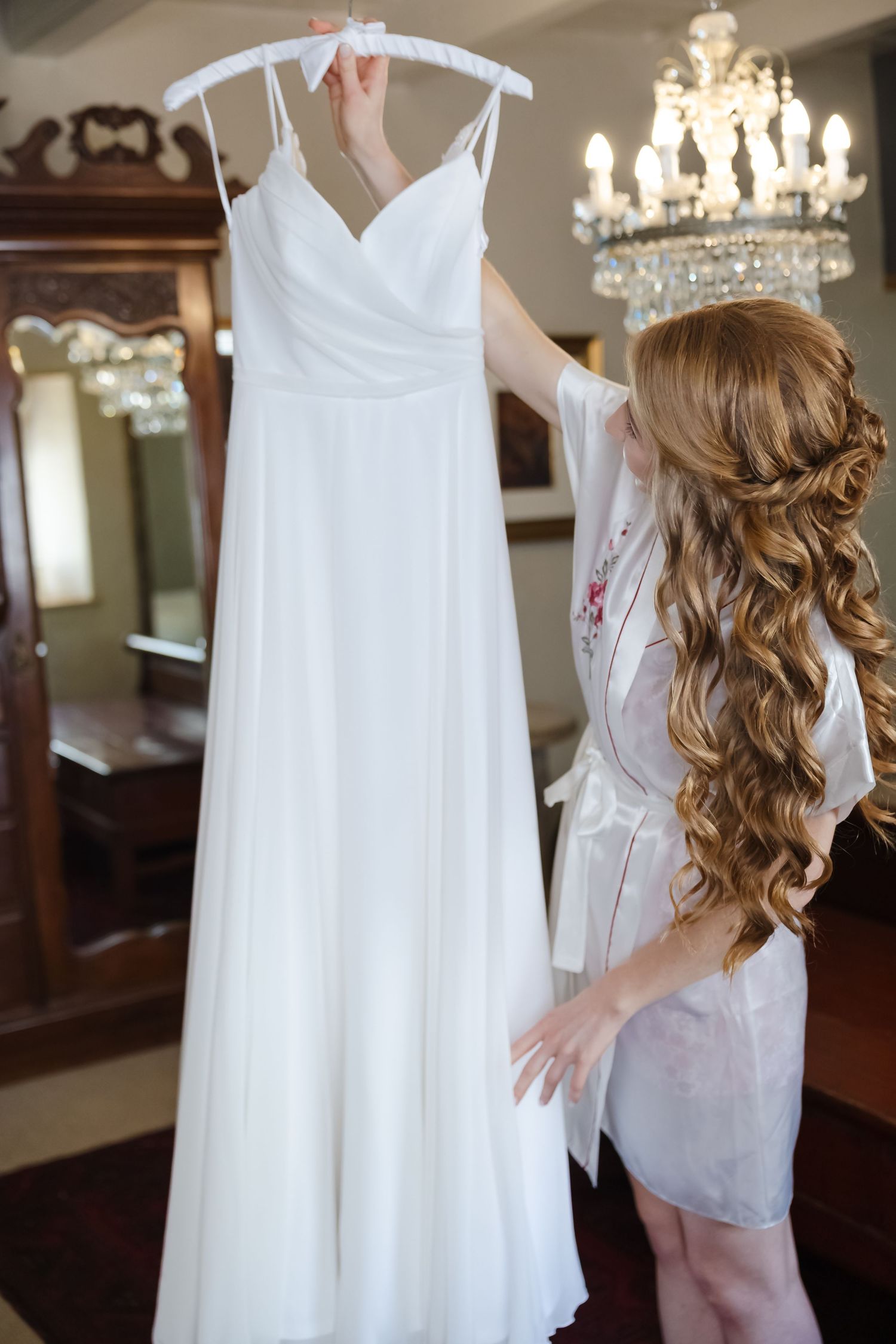 A flowing white wedding dress hangs on display as someone in a silk robe with long curled hair admires it under a chandelier.