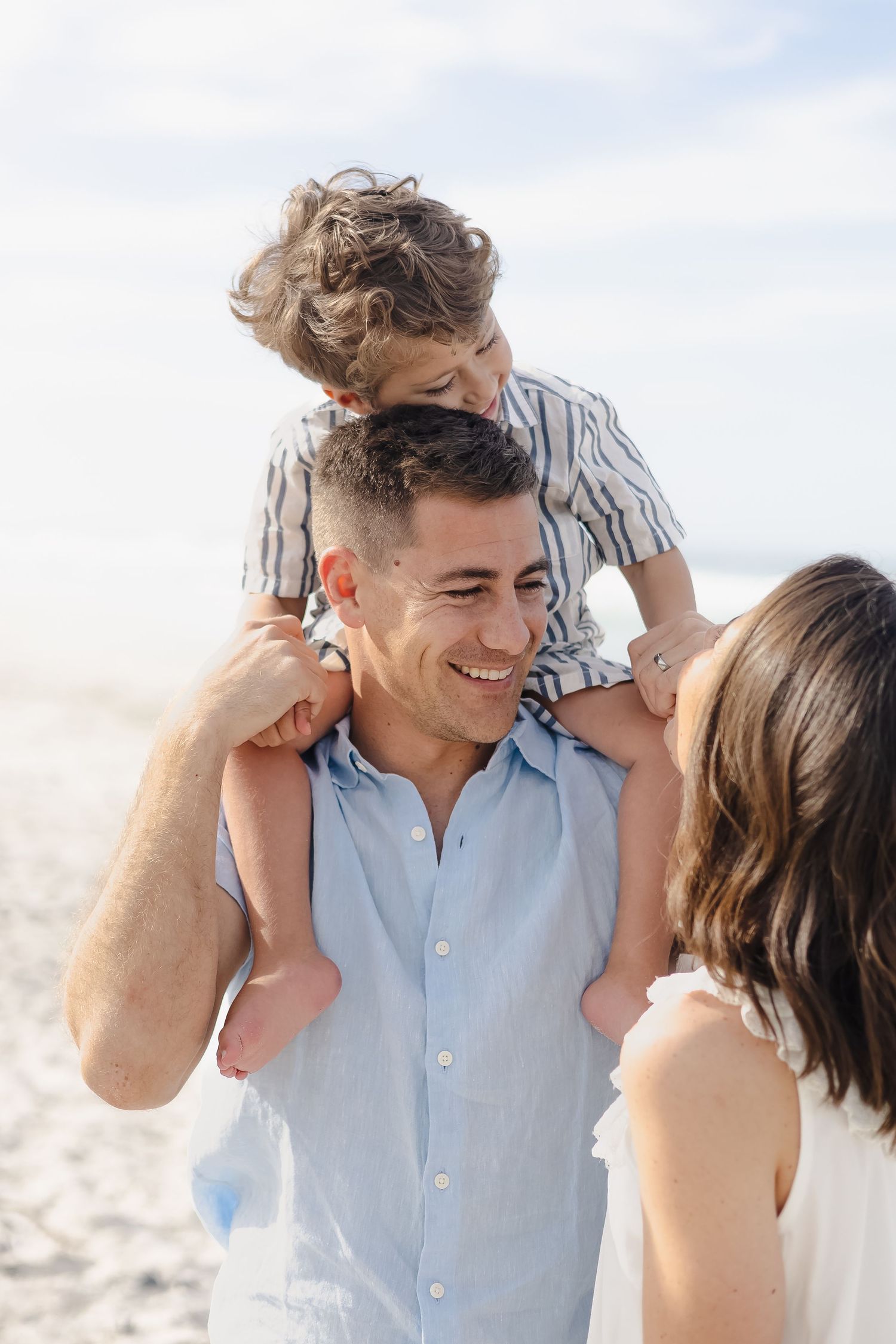 A happy family at the beach, with a smiling man carrying a young child on his shoulders beside a woman.
