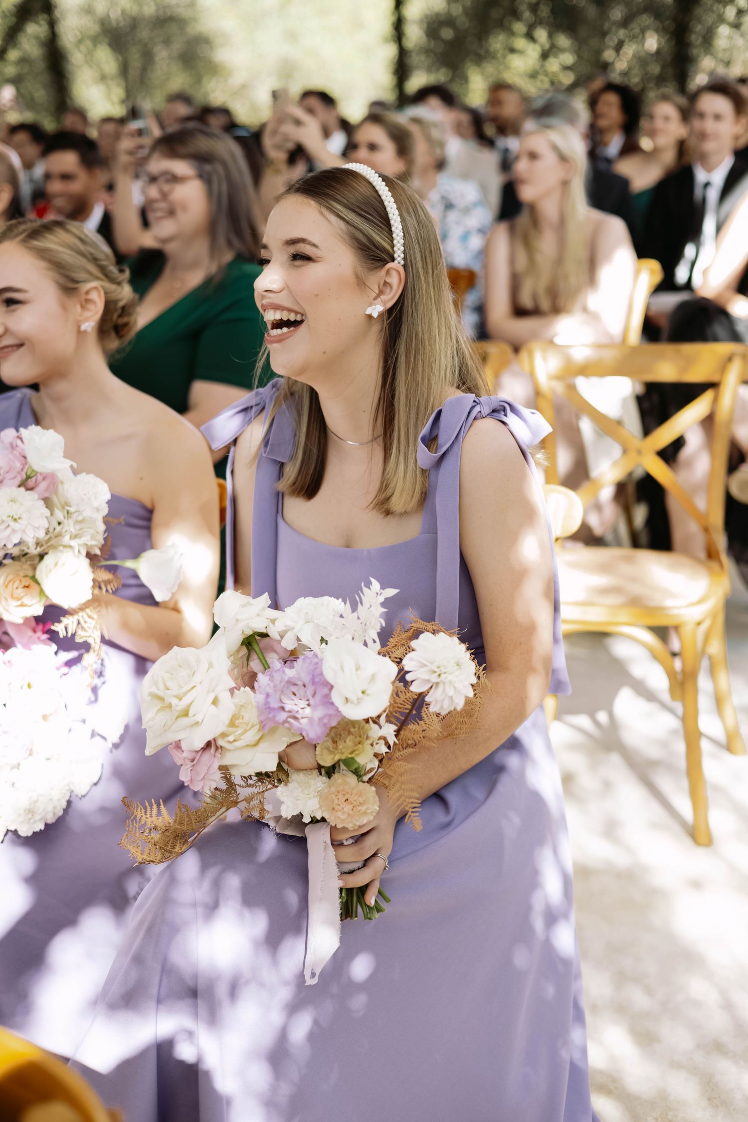 Bridesmaids in lavender dresses laugh joyfully while holding pastel bouquets during an outdoor wedding ceremony.