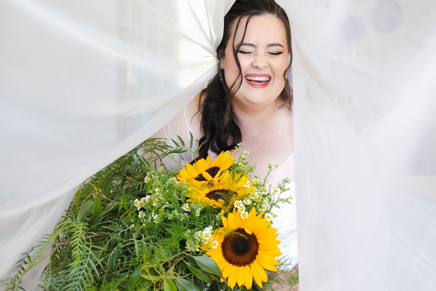 Laughing bride with sunflower bouquet peeking through white veil on her wedding day.
