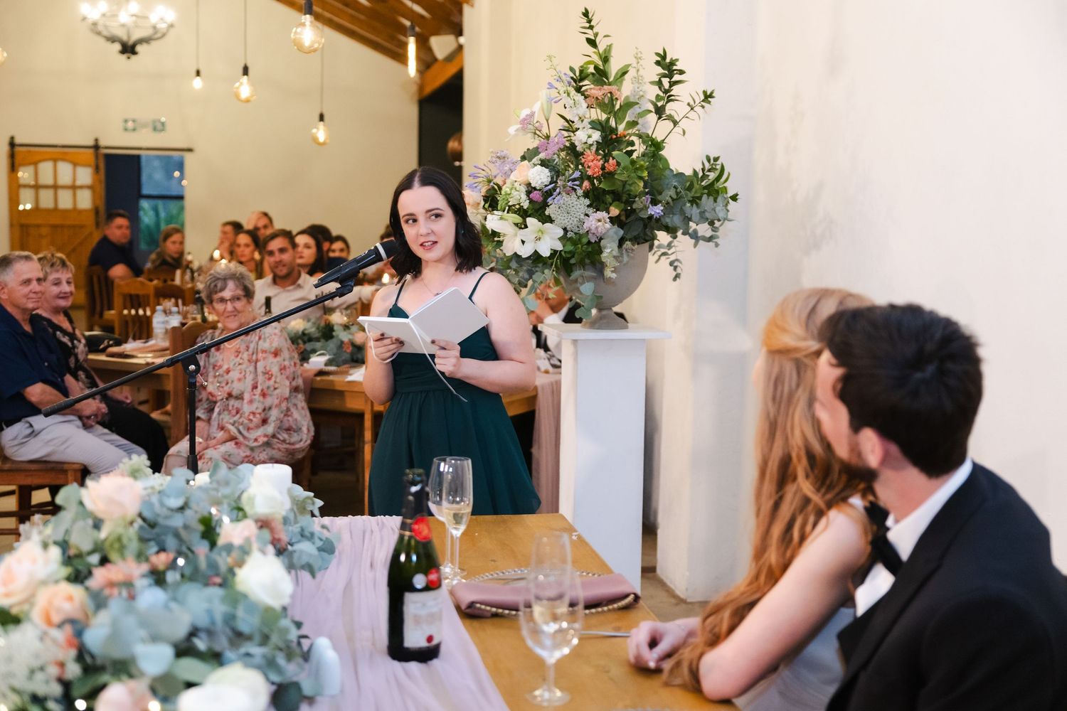 A wedding reception dinner with elegant floral centerpieces and guests gathered around tables in a rustic venue.