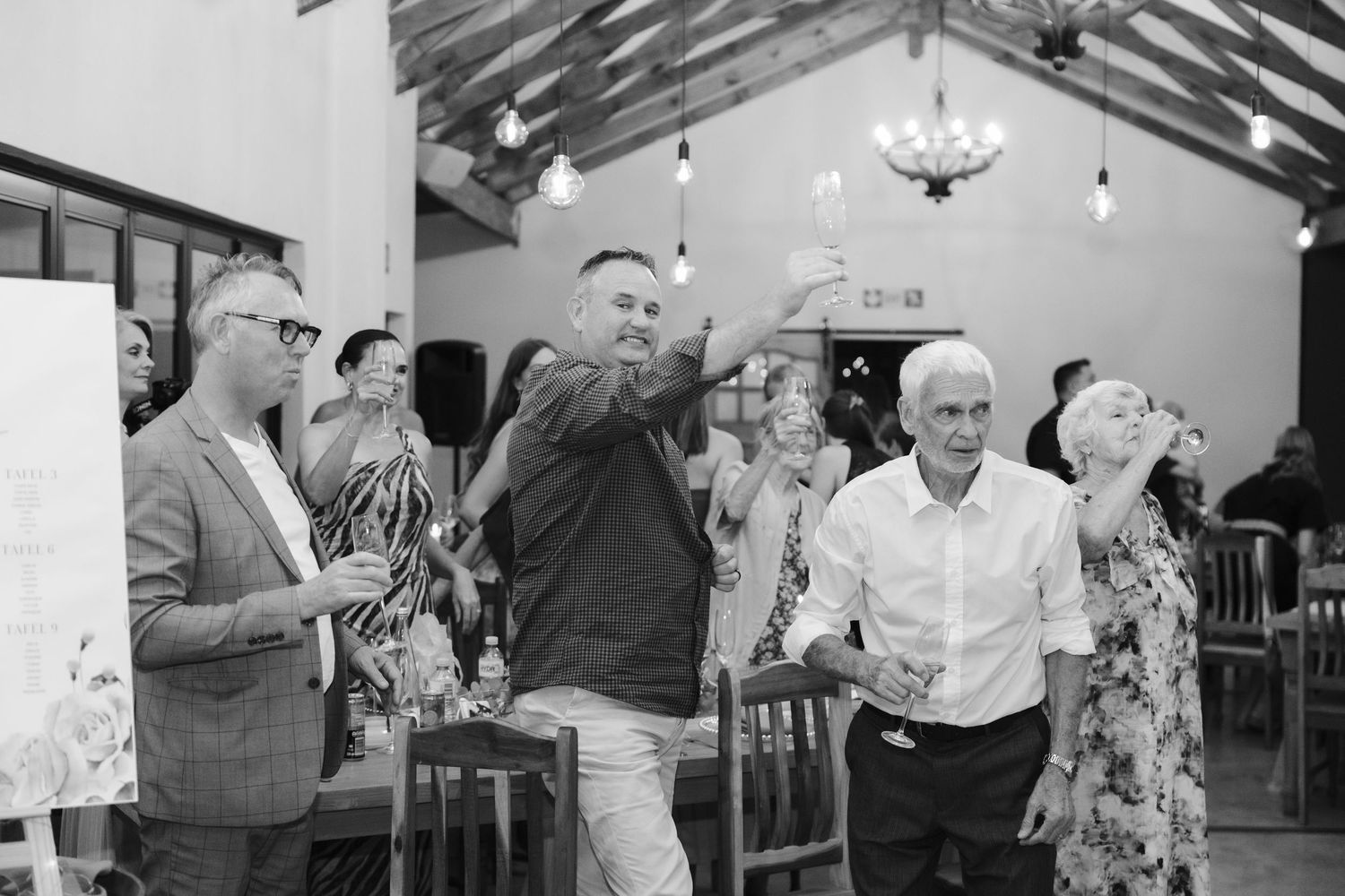 Wedding guests dancing and celebrating together in a rustic venue with exposed wooden beams and chandeliers.