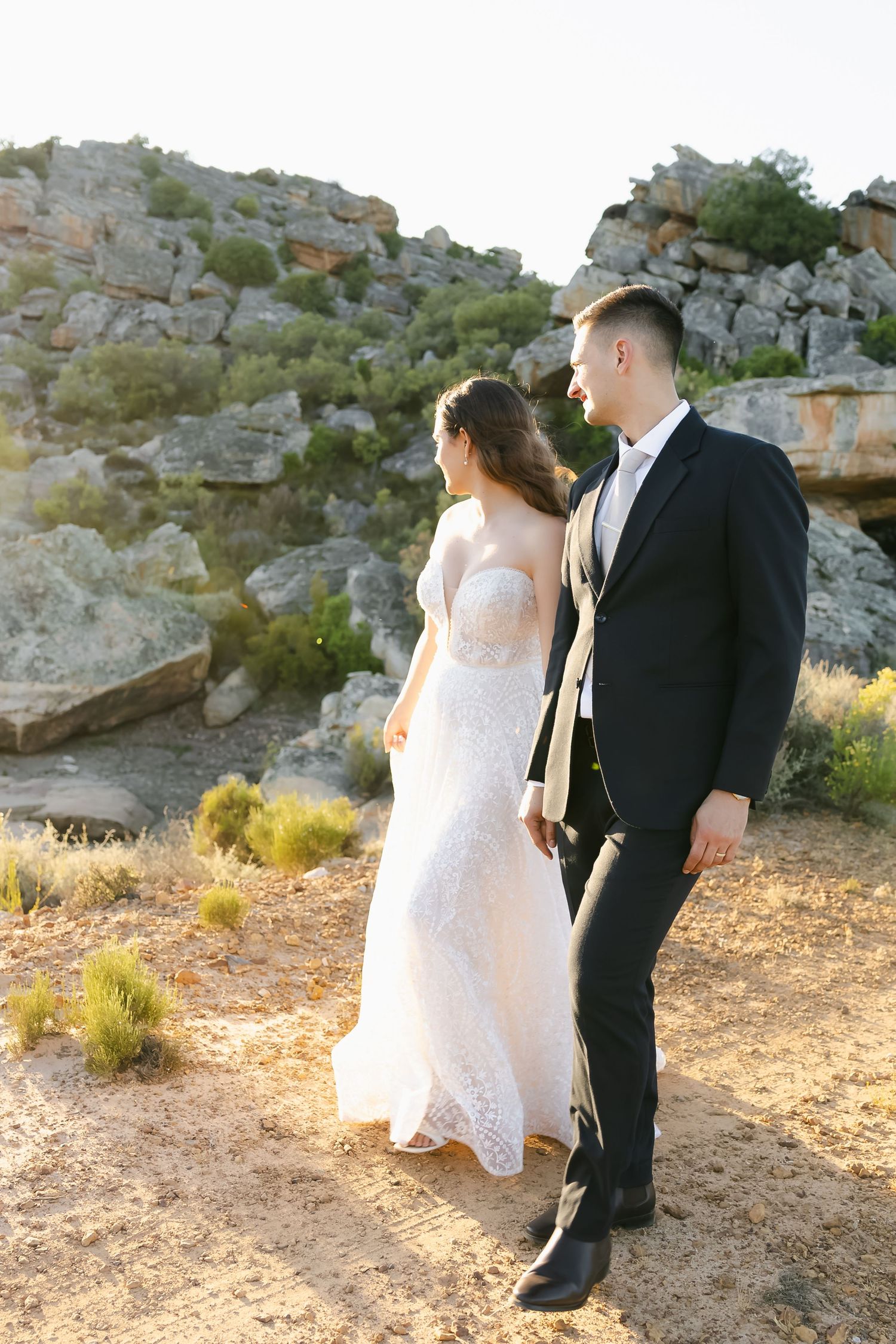A wedding couple poses together on rocky terrain during golden hour in formal attire.