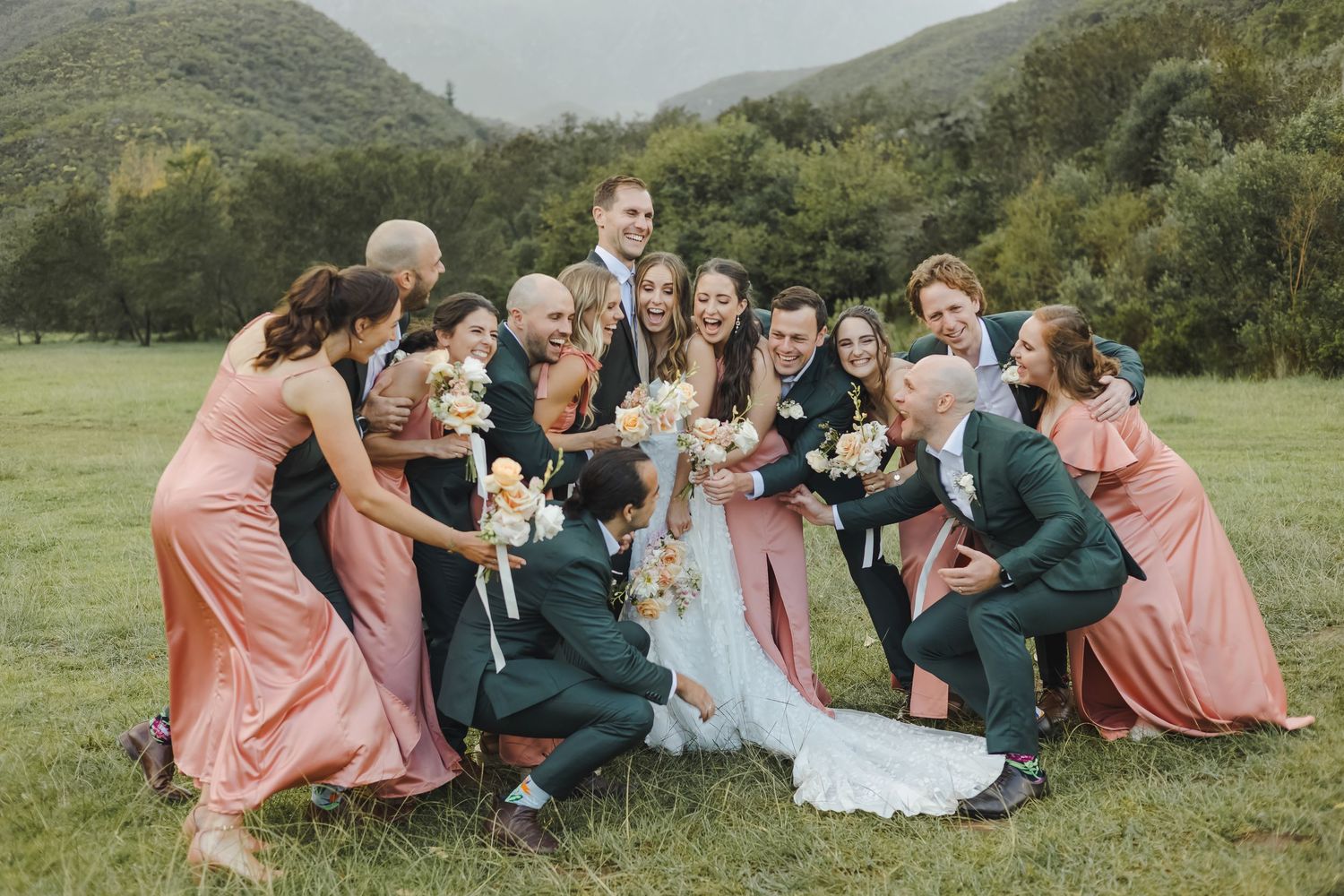 Wedding party relaxing on grass with mountain scenery in the background.