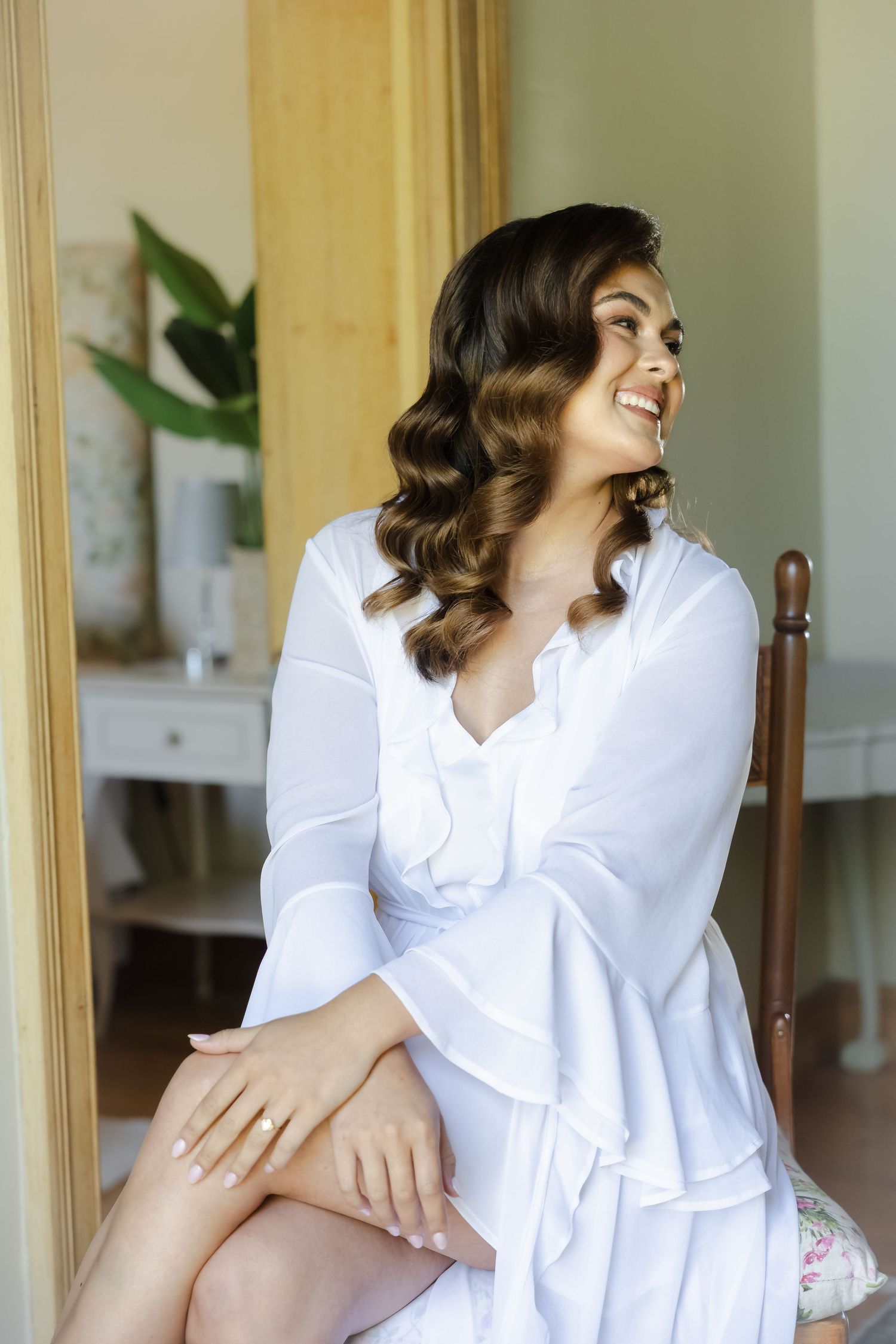 Woman in white robe sitting and smiling while getting ready in a sunlit room with wooden furniture.