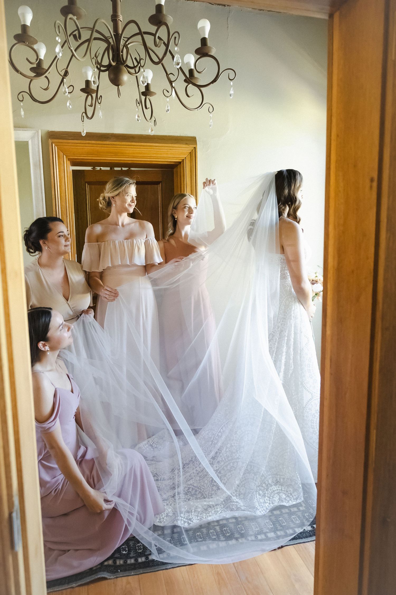 Bridesmaids in pink dresses help adjust a flowing white wedding veil in an elegant room with a chandelier.