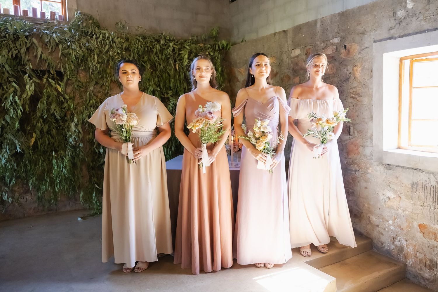 Bridesmaids in blush and neutral tones hold bouquets while standing together in a rustic stone-walled room.