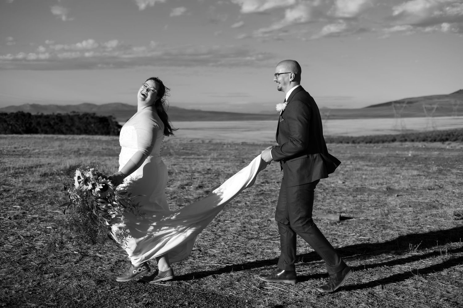 Black and white photo of a laughing bride holding flowers and groom walking hand in hand in an open field.