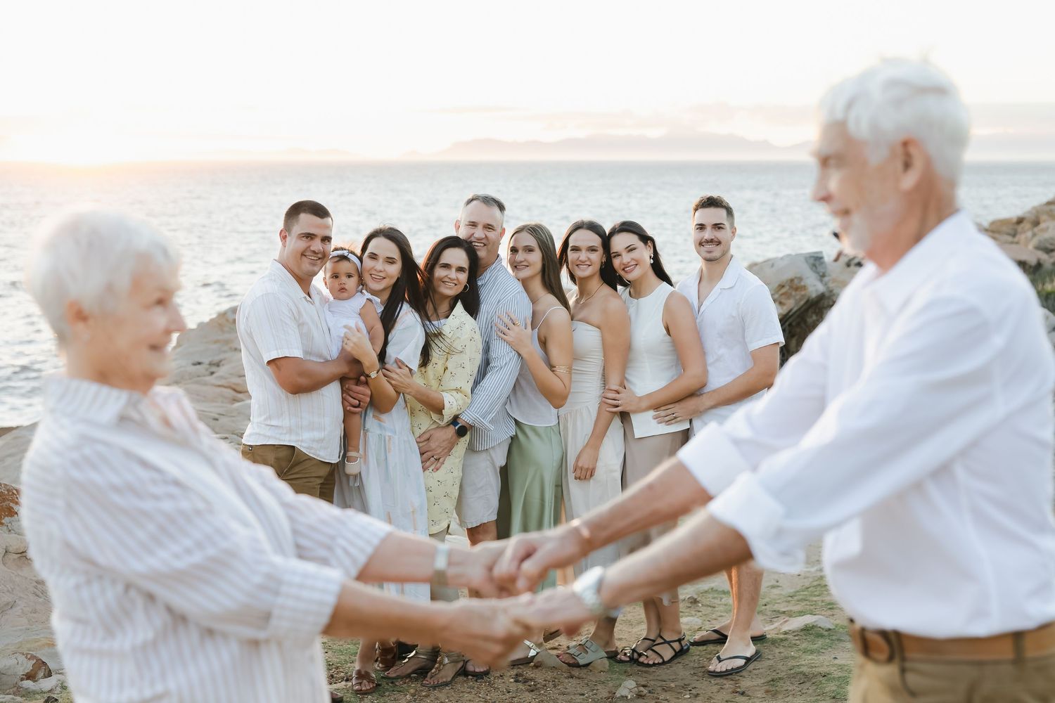Large family gathering on a rocky beach at sunset, holding hands and smiling together near the ocean.