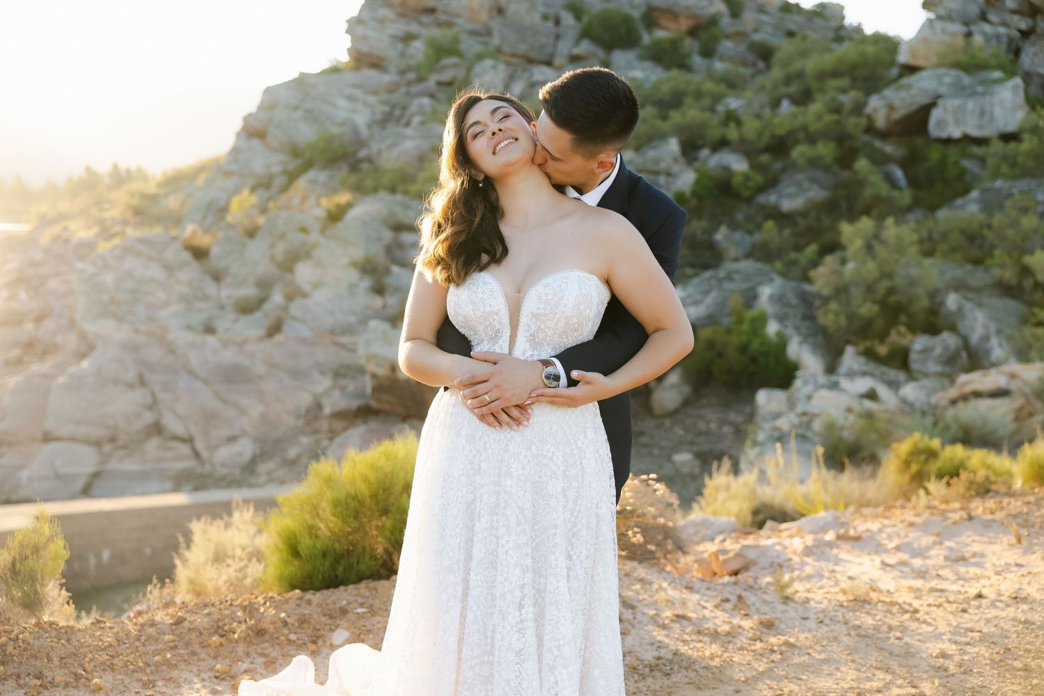 Wedding couple posing together on rocky terrain with mountains and desert landscape in the background.