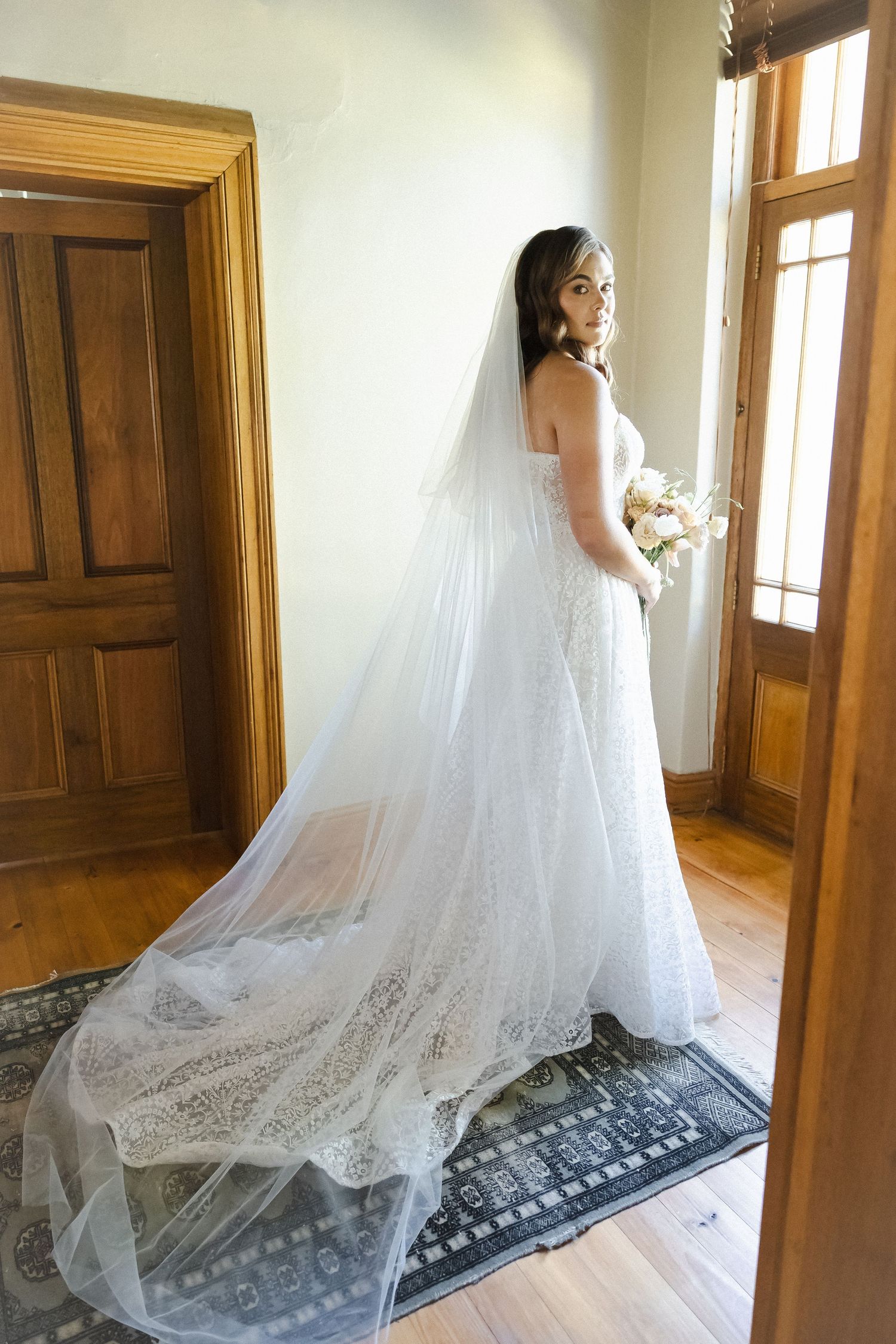 A bride in a flowing white wedding gown and long veil stands elegantly in a sunlit hallway with wooden doors.