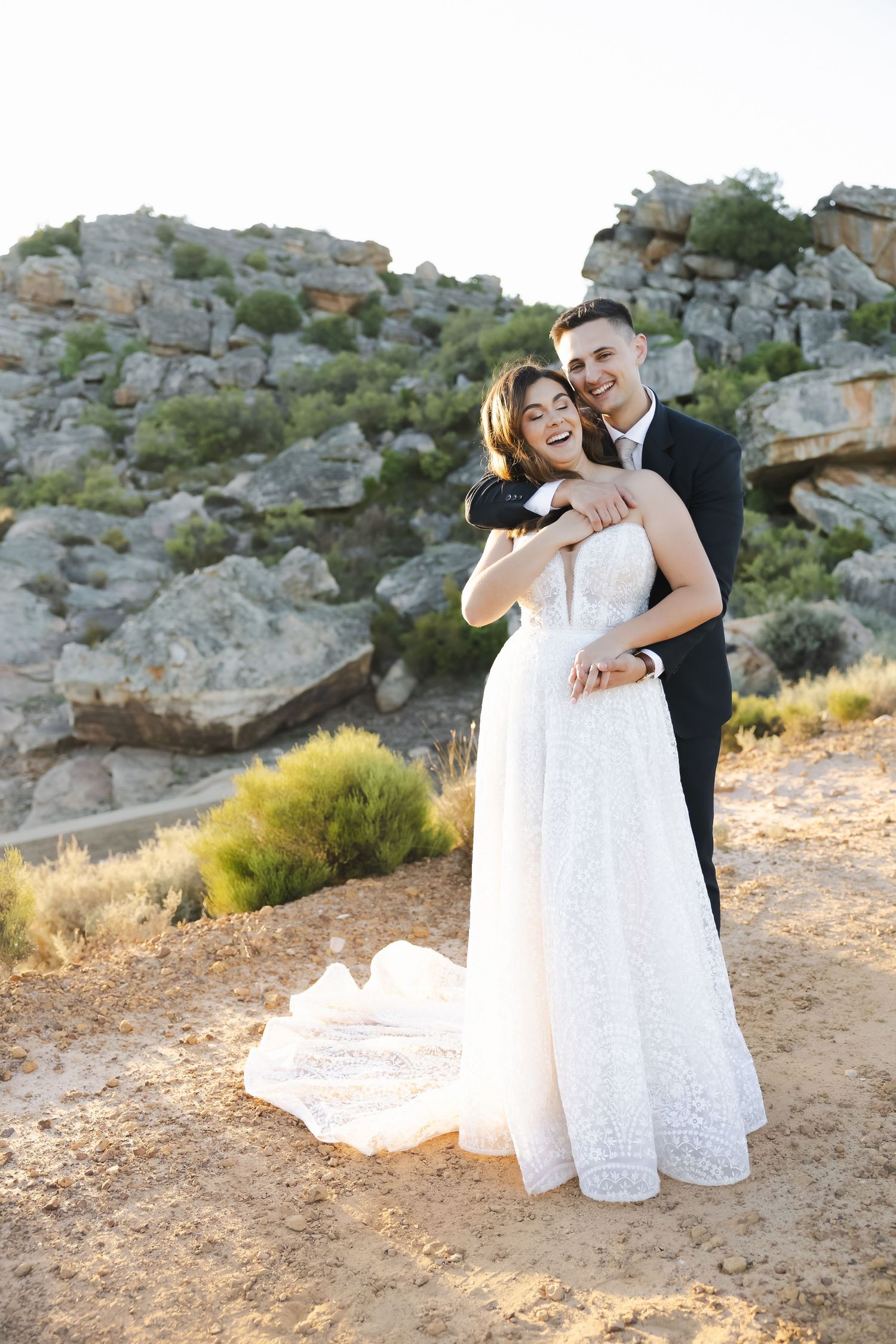 Joyful wedding couple laughing and embracing on rocky desert terrain during a sunset photoshoot.