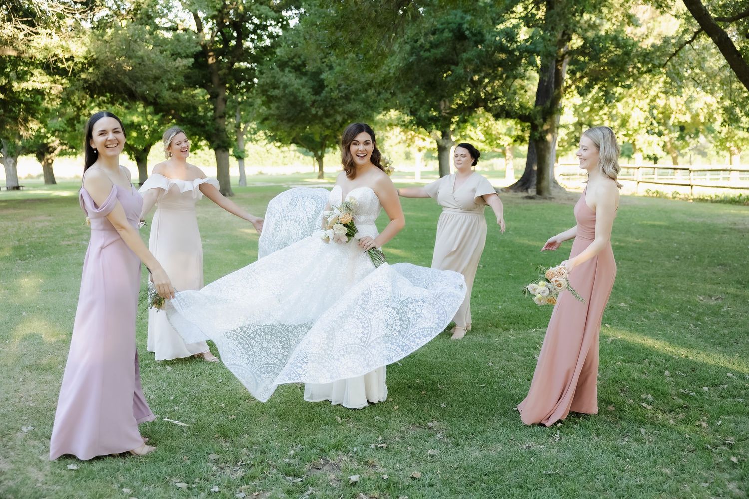 Bridesmaids in blush pink dresses walk together through a sunlit garden setting during a summer wedding celebration.