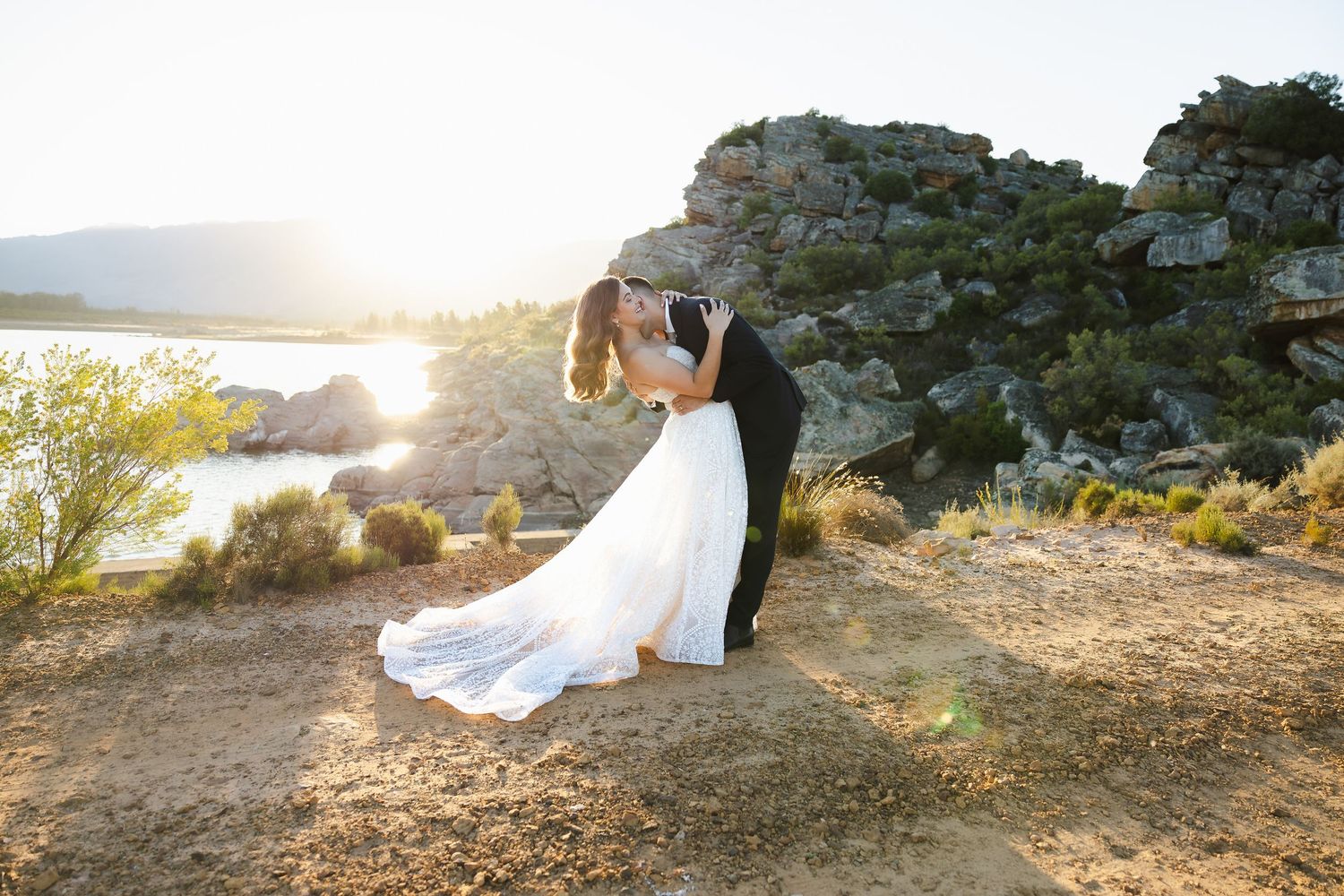A couple in wedding attire poses on a rocky cliff overlooking the ocean during golden hour sunset.