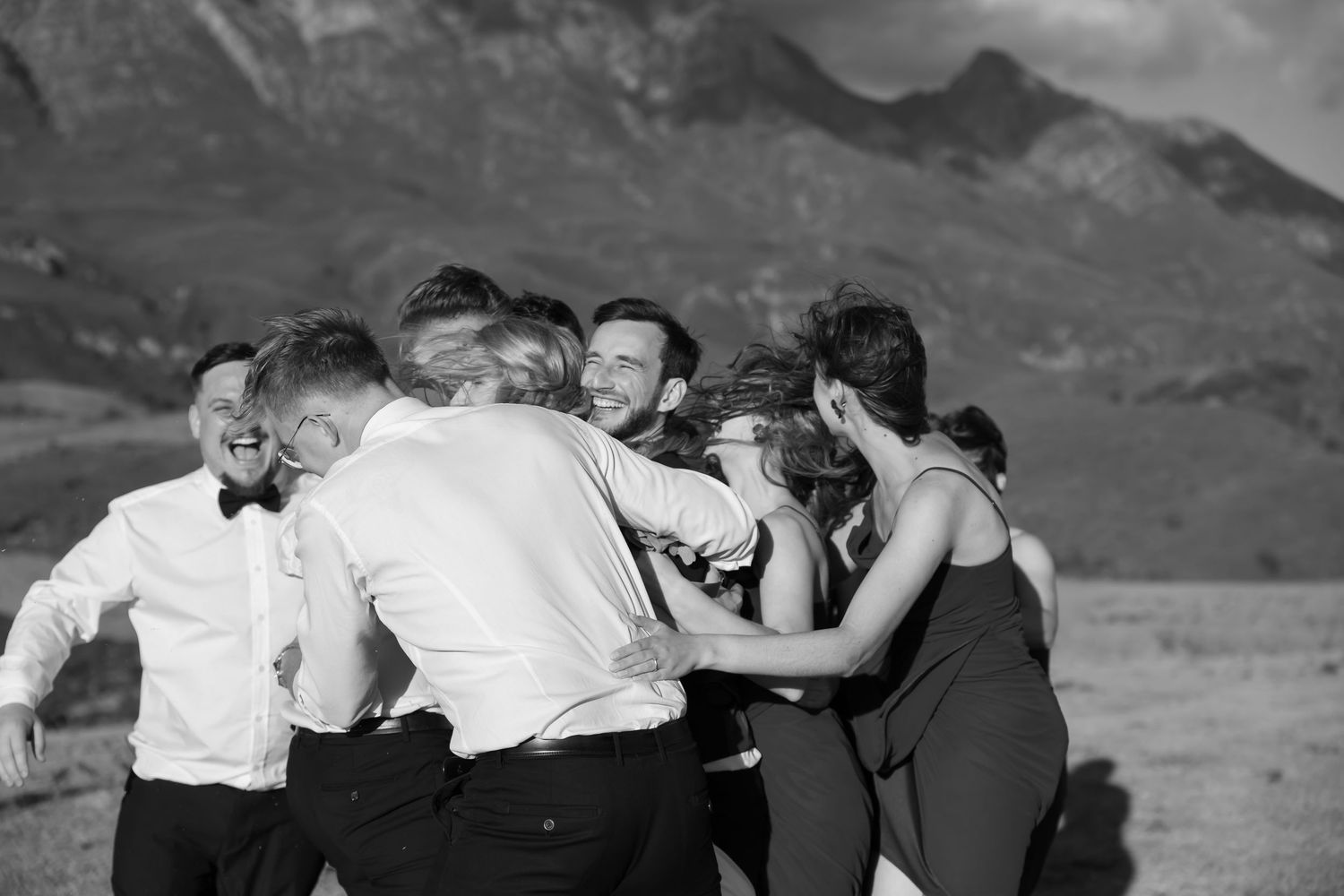 A group of people dancing and celebrating in black and white formal attire against a mountain landscape backdrop.