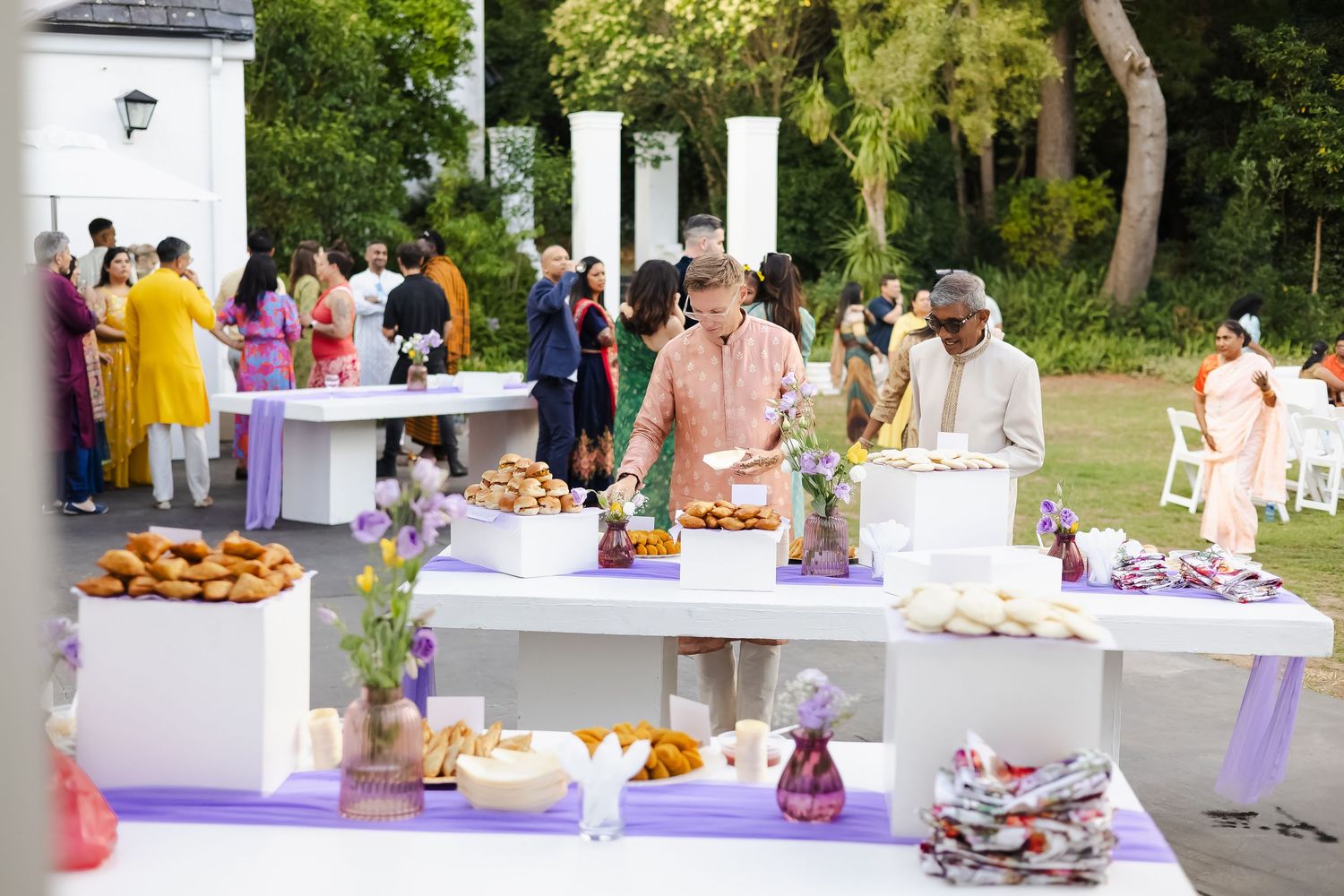 Outdoor wedding reception setup with white tables, colorful decorations and refreshments under trees.
