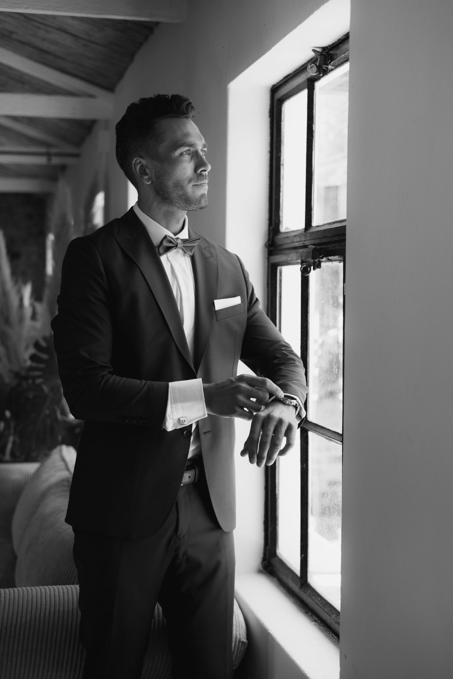 Black and white photo of groom getting ready by a window in formal attire.