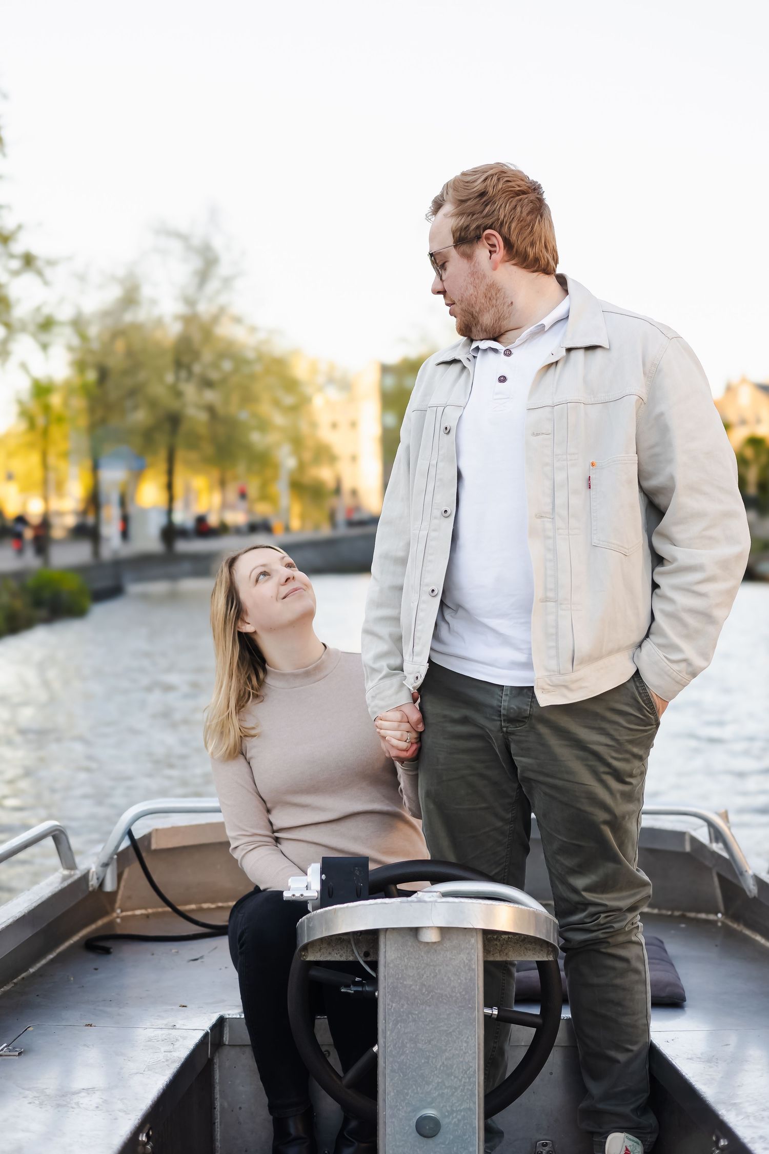 Couple holding hands on a boat by an Amsterdam canal, woman sitting looking up at standing man in beige jacket.