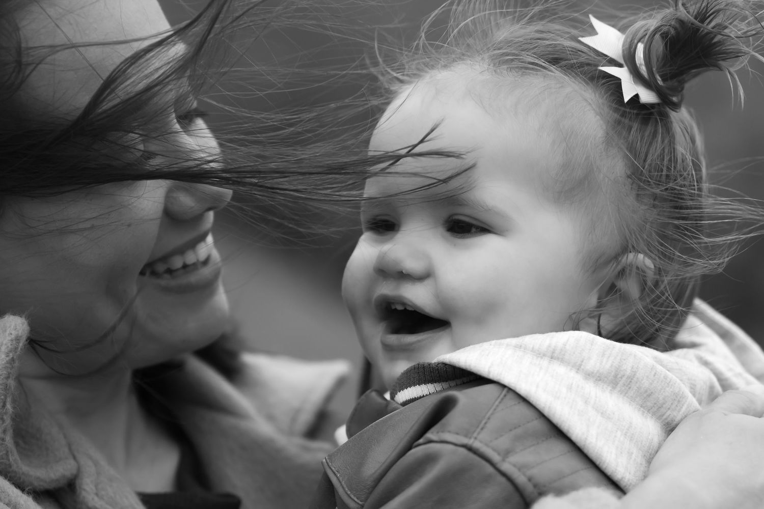 Black and white photo of a smiling mother holding a laughing baby girl with a bow in her hair.