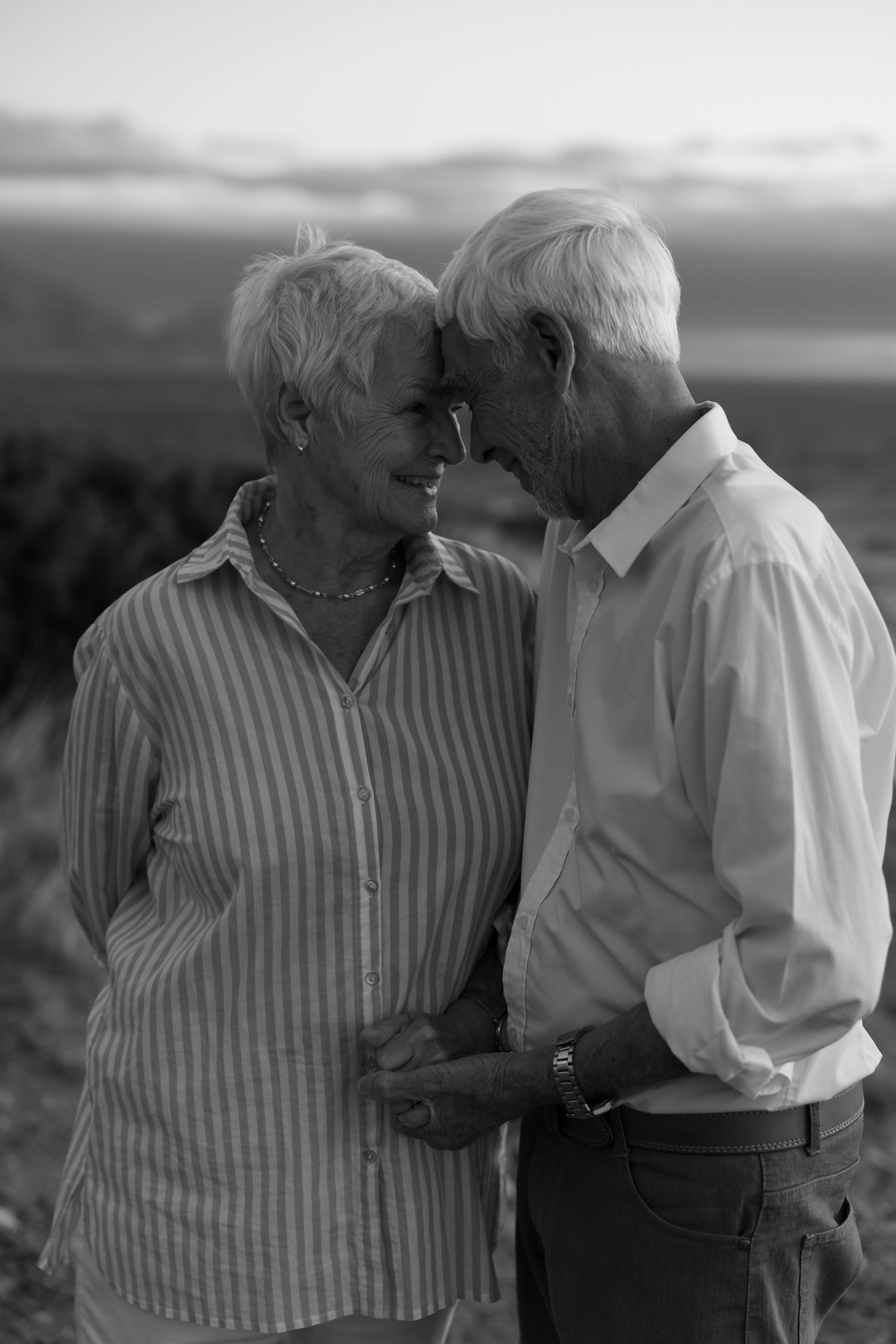 Black and white photo of an elderly couple tenderly touching foreheads outdoors at sunset.