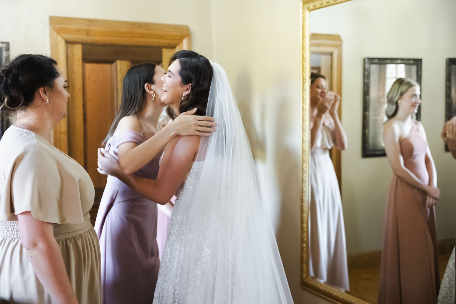 Bride in white wedding gown and veil gets ready with bridesmaids in mauve dresses while looking in mirror.