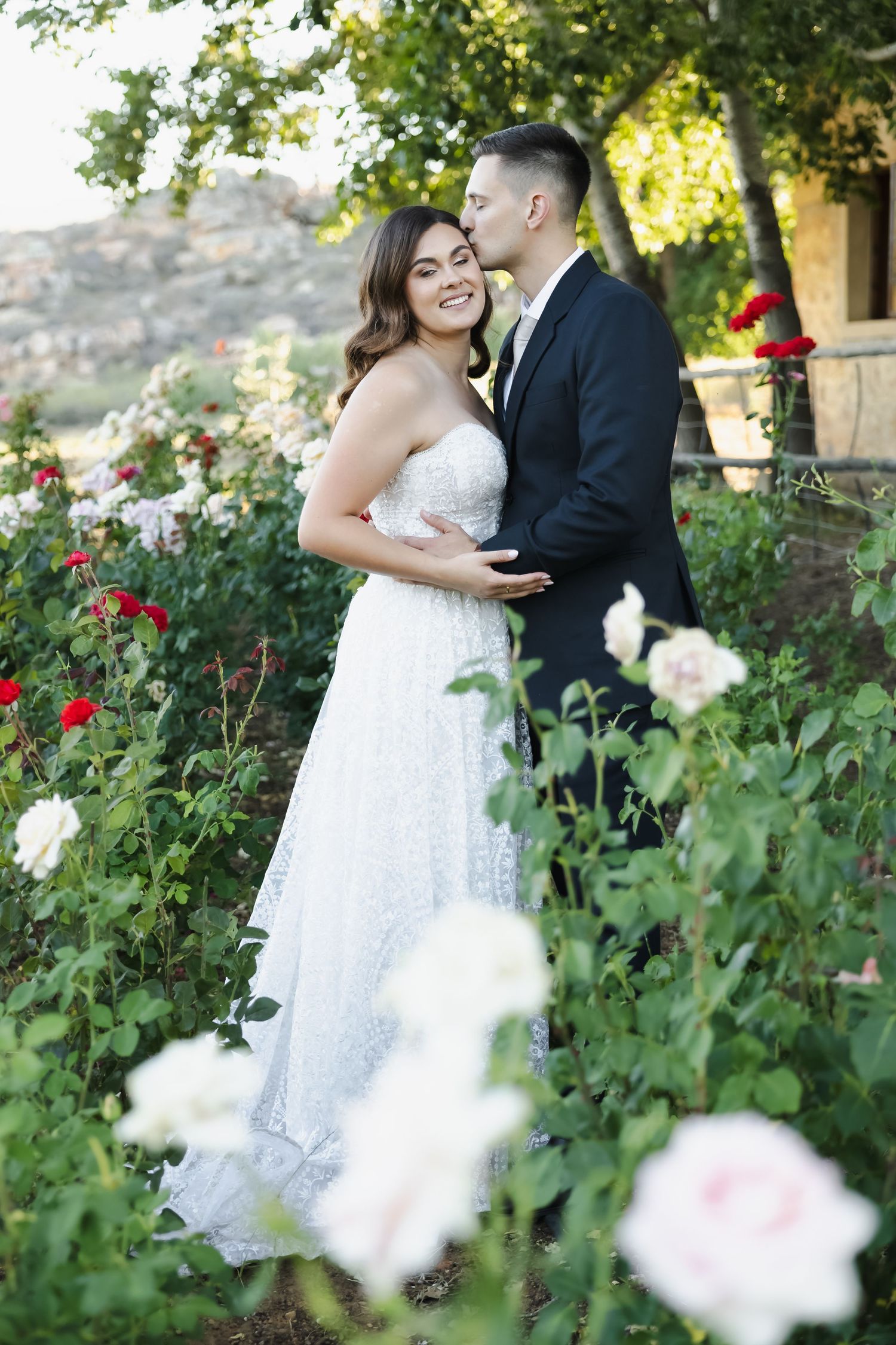 A romantic wedding couple embraces in a garden with blooming red roses and lush greenery.