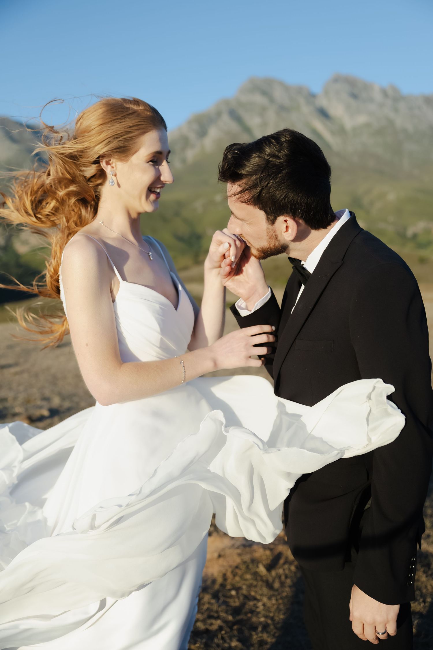 A couple in wedding attire dances playfully against a mountainous backdrop with the bride's white dress flowing in the wind.
