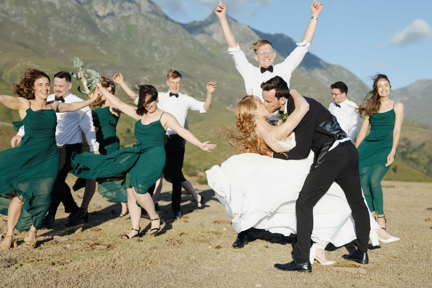 Bridal party performs synchronized dance moves on a mountainside during a wedding photoshoot.