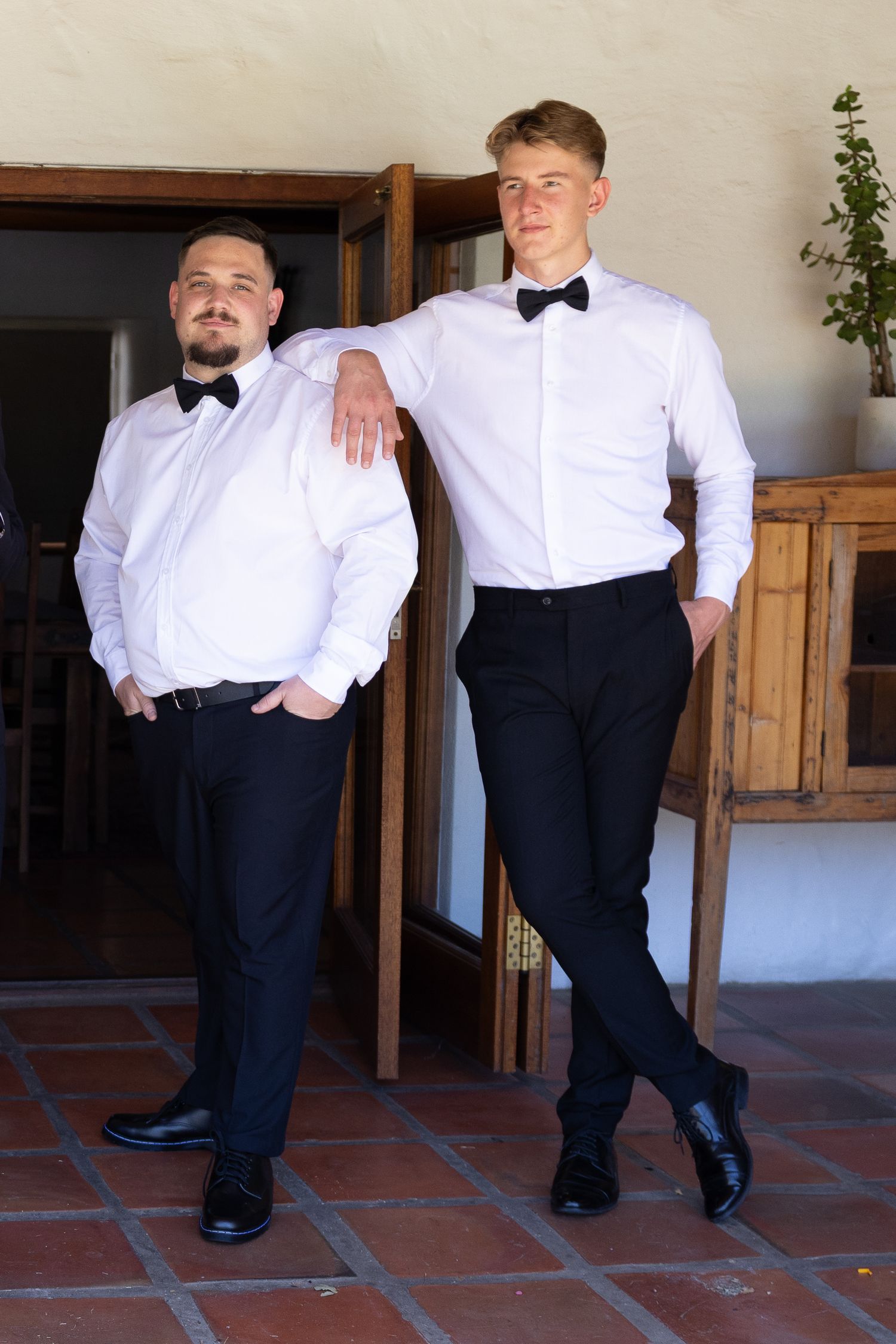 Two people in formal white shirts and bowties pose together against a light colored wall.
