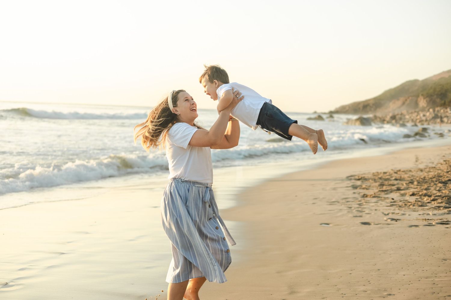 Mother joyfully lifting her young son on a sunny beach with ocean waves in the background.