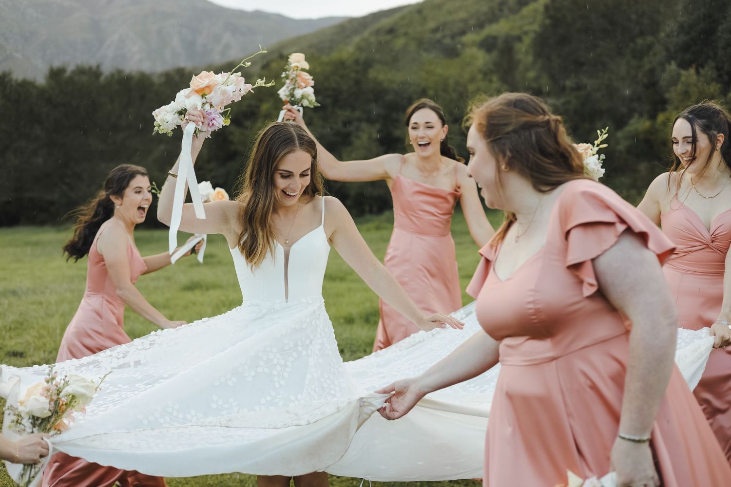 A bridal party in coral dresses spins and dances outdoors with the bride in a flowing white gown against mountain scenery.