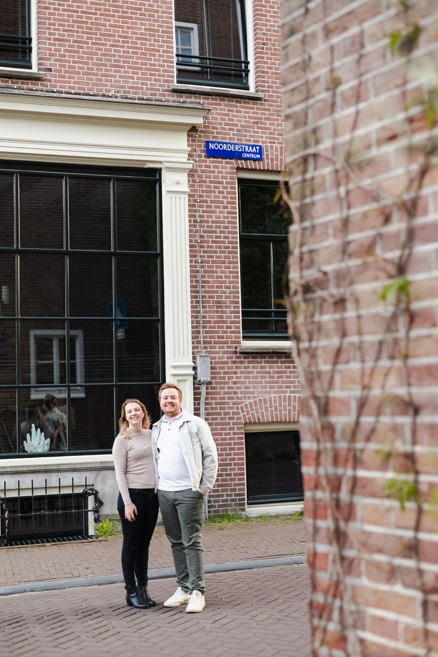 Smiling couple standing together on a brick street in Amsterdam near a Noorderstraat sign.