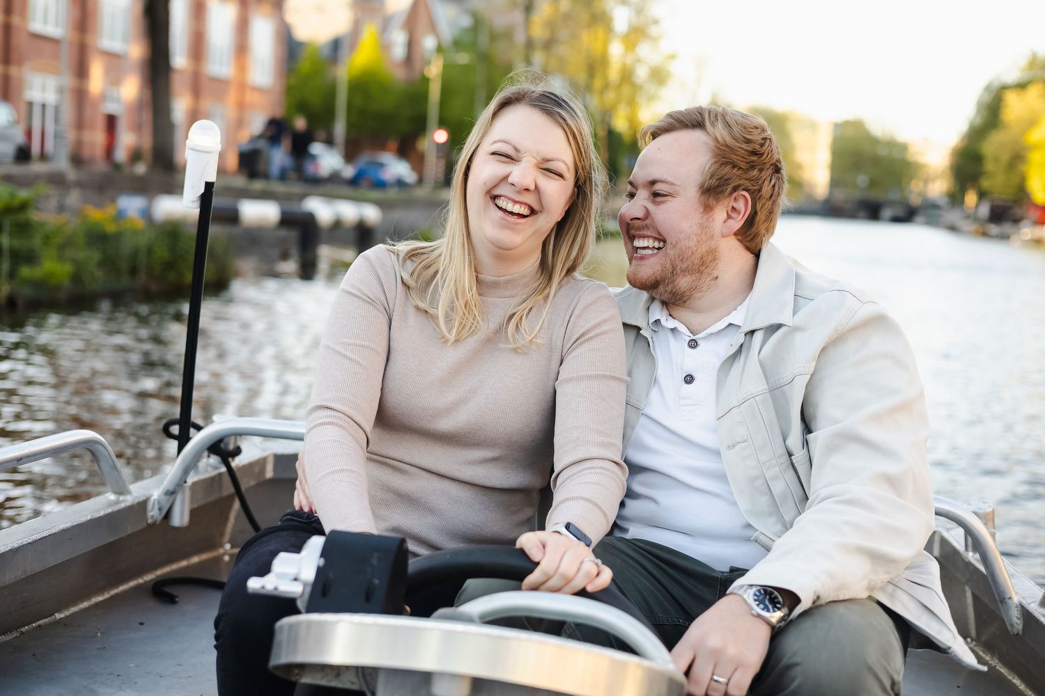 Couple laughing joyfully on a boat along a scenic canal, enjoying a sunny day outdoors together.