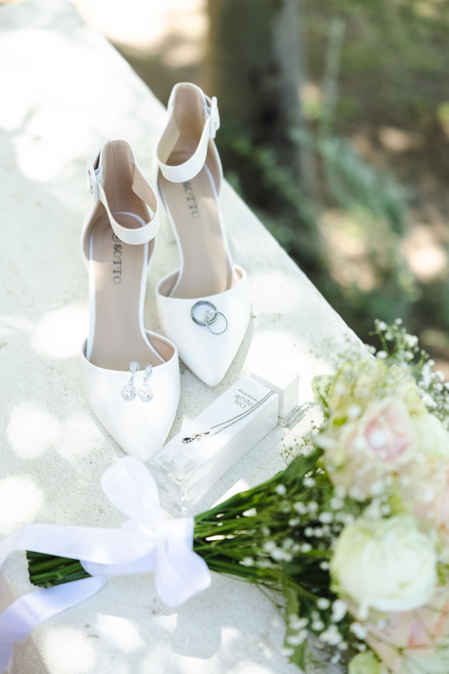 Elegant white bridal shoes with wedding rings displayed alongside delicate white floral arrangements and greenery.