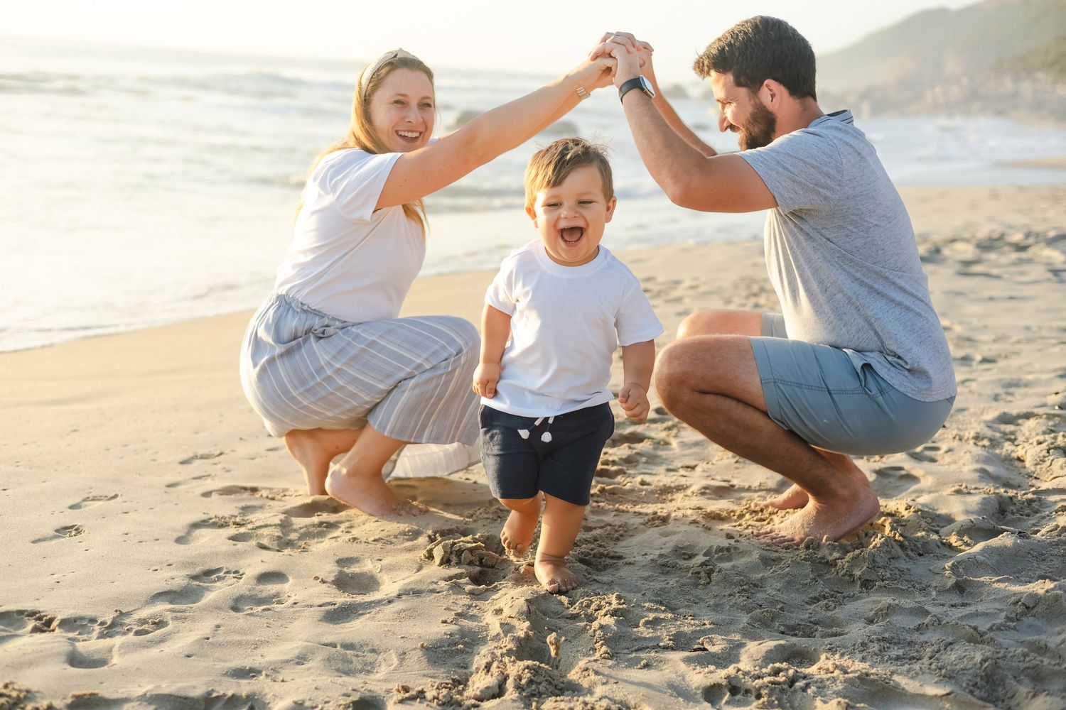 A happy young family with a toddler playing on a sunny beach, parents holding hands above child's head.