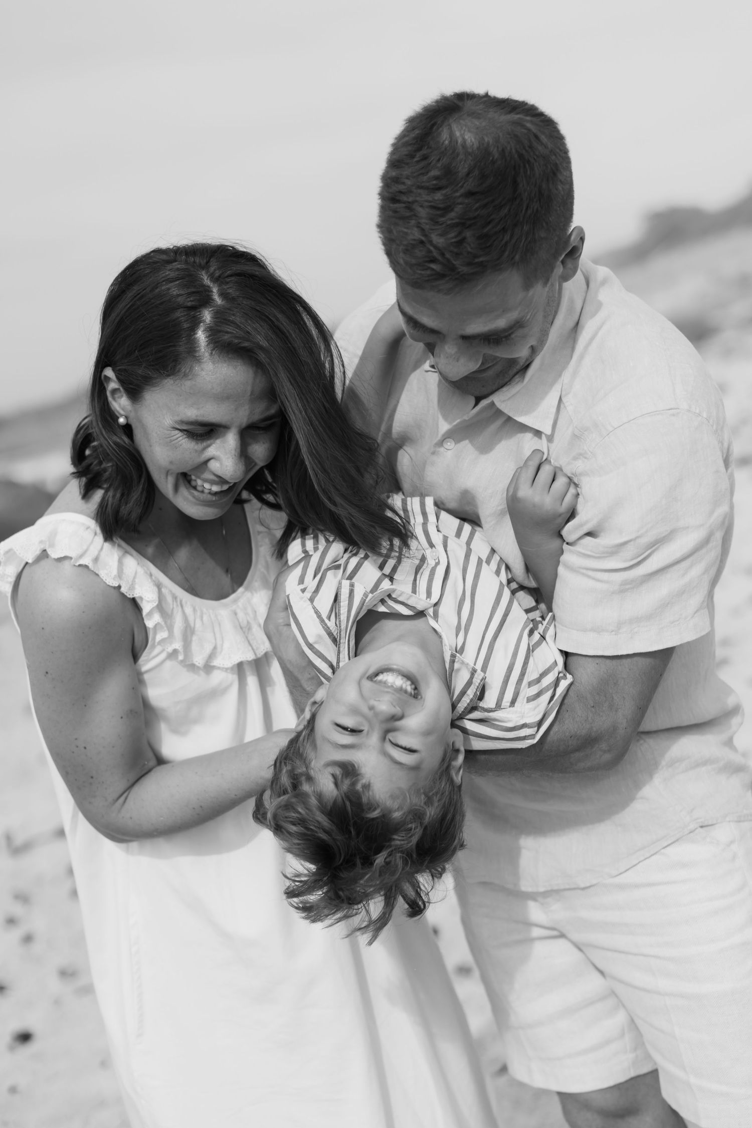 Black and white photo of a joyful family at the beach, parents laughing as they flip their child upside down.