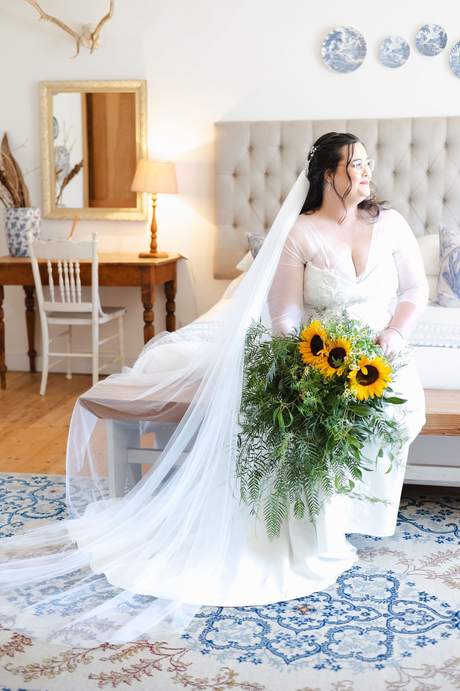 Bride in white gown with long veil holds sunflower bouquet in elegant bedroom.