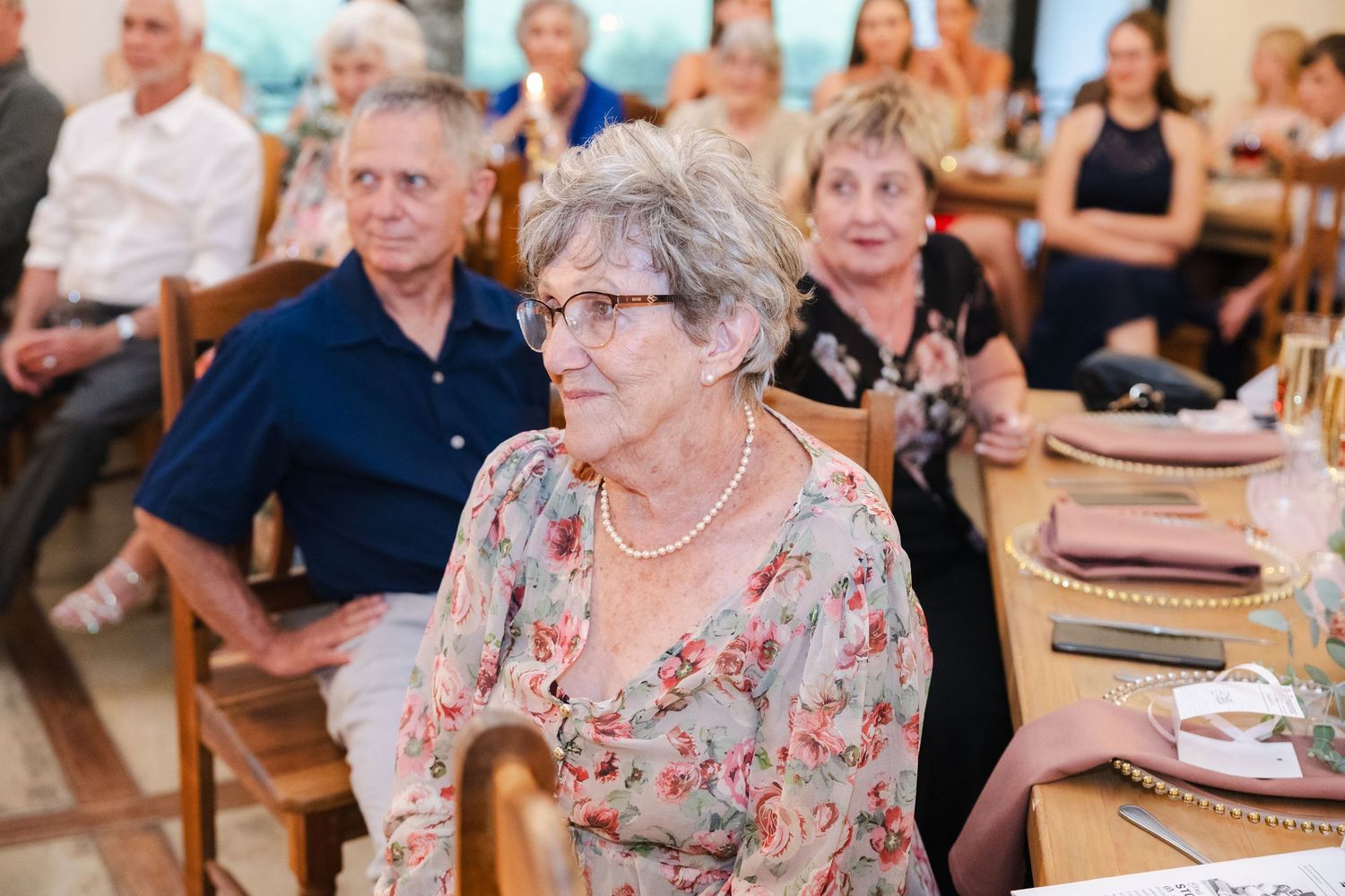 Elderly people sitting at tables during a social gathering or event with blue balloons in the background.