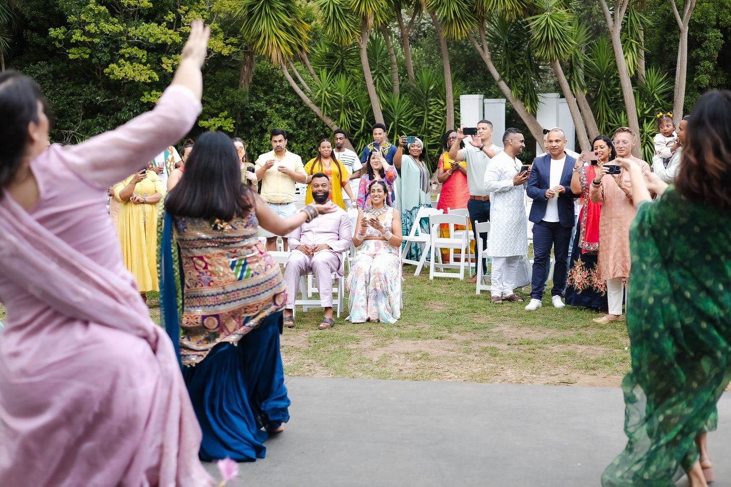 A crowd of people in colorful outfits dance in a circle on a sunny outdoor lawn during a festive celebration.
