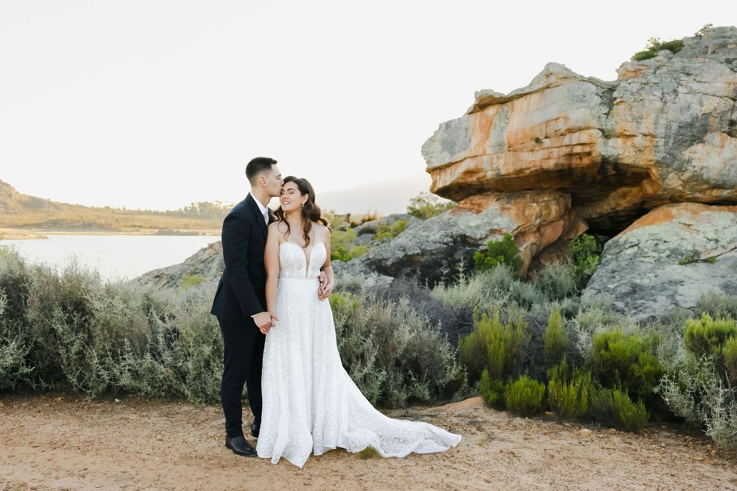 A couple in wedding attire embraces in front of a dramatic rock formation in a desert landscape at sunset.