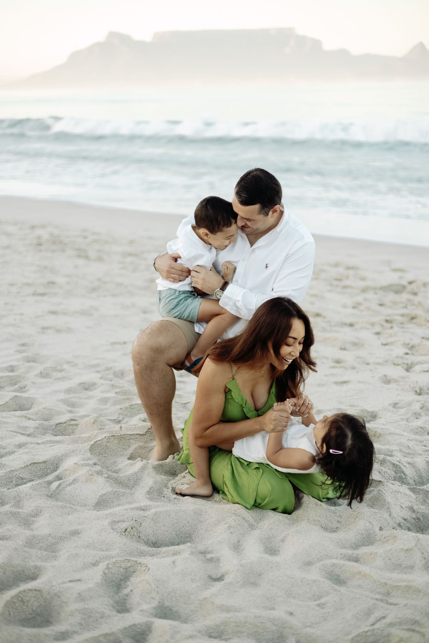 Intimate family moment captured on a sandy beach with ocean waves in the background.